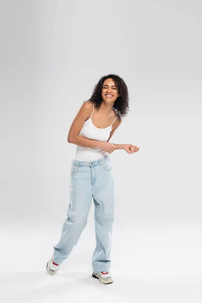 Longitud completa de la alegre mujer afroamericana en pantalones vaqueros azules y zapatillas de deporte bailando sobre fondo gris - foto de stock