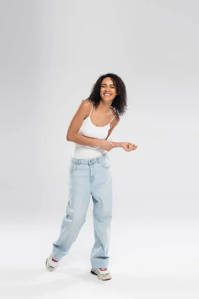 Comprimento total de mulher americana africana alegre em jeans azuis e tênis dançando em fundo cinza — Fotografia de Stock