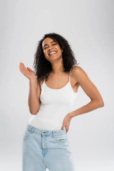 Joyful african american woman in white tank top waving hand and smiling isolated on grey — Stock Photo