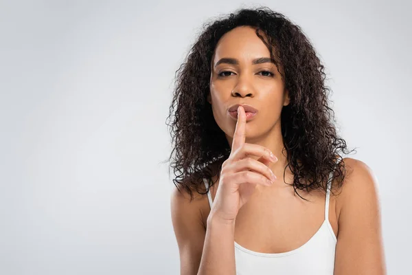 Young african american woman with wavy brunette hair showing looking at camera and showing hush sign isolated on grey — Stock Photo