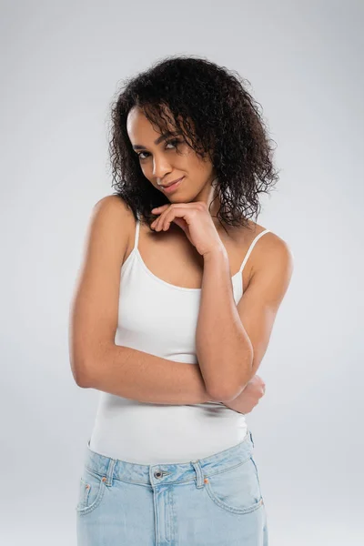 Coqueta y sonriente mujer afroamericana en camiseta blanca sostenida de la mano cerca de la cara y mirando a la cámara aislada en gris - foto de stock