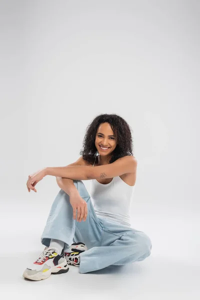 Pleine longueur de femme américaine africaine joyeuse avec des cheveux bruns bouclés assis en jeans bleu sur fond gris — Photo de stock
