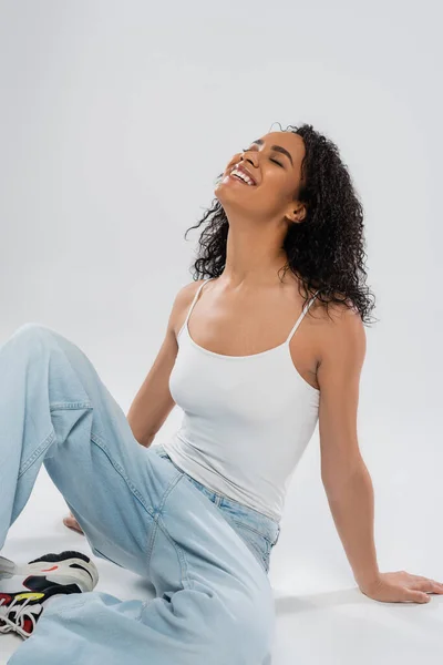 Joven y feliz mujer afroamericana en jeans sonriendo con los ojos cerrados mientras se sienta sobre fondo gris - foto de stock
