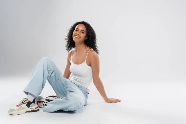 Full length of stylish african american woman in jeans and tank top sitting and looking at camera on grey background — Stock Photo
