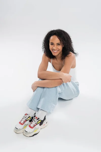 Longitud completa de la alegre mujer afroamericana en zapatillas de deporte y jeans sentado sobre fondo gris - foto de stock