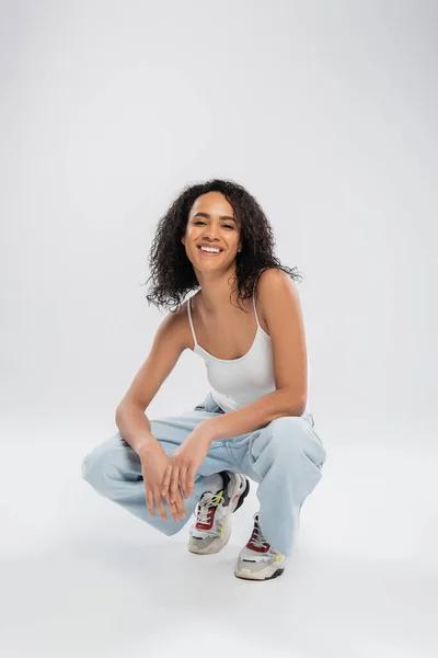 Full length of cheerful african american woman in blue jeans posing on haunches and smiling at camera on grey background — Stock Photo