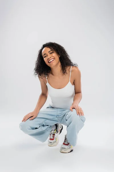 Full length of overjoyed african american woman in jeans and sneakers sitting on haunches on grey background — Stock Photo