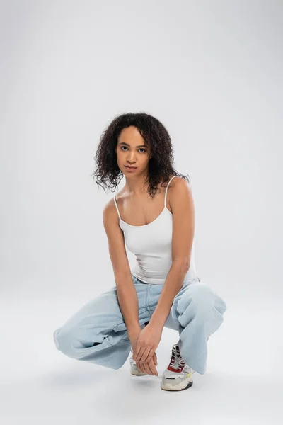 Full length of african american woman in white tank top and jeans with sneakers sitting on haunches on grey background — Stock Photo