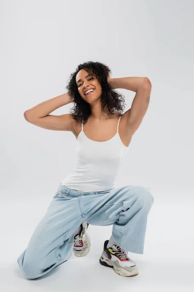 Piena lunghezza di donna afroamericana sorridente in canotta bianca e jeans toccare i capelli ondulati su sfondo grigio — Foto stock