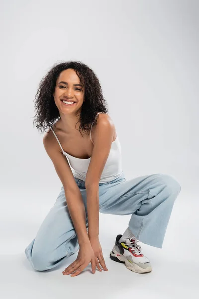 Full length of excited african american woman in tank top and blue jeans posing on grey background — Stock Photo