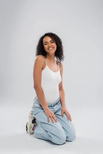 Full length of pretty african american woman in jeans and white tank top sitting on haunches on grey background — Stock Photo