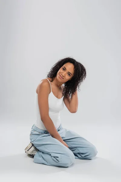 Full length of positive african american woman in tank top and jeans touching hair while sitting on grey background — Stock Photo
