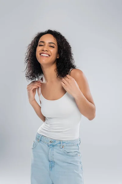 Mujer afroamericana llena de alegría en camiseta blanca sonriendo a la cámara aislada en gris - foto de stock