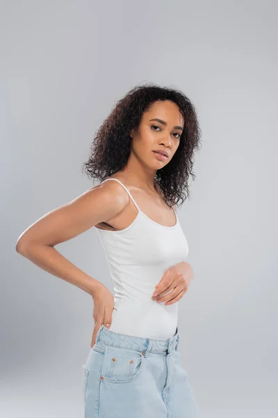 Brunette african american woman in white tank top holding hand behind back while looking at camera isolated on grey — Stock Photo