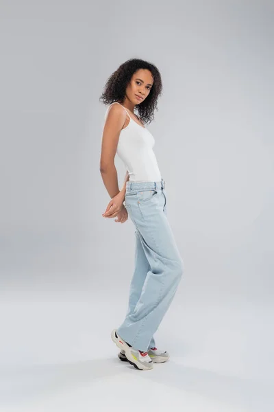 Pleine longueur de mince femme afro-américaine en jeans bleus et débardeur blanc sur fond gris — Photo de stock