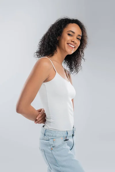 Morena mujer afroamericana en camiseta blanca posando con las manos detrás de la espalda y sonriendo a la cámara aislada en gris - foto de stock