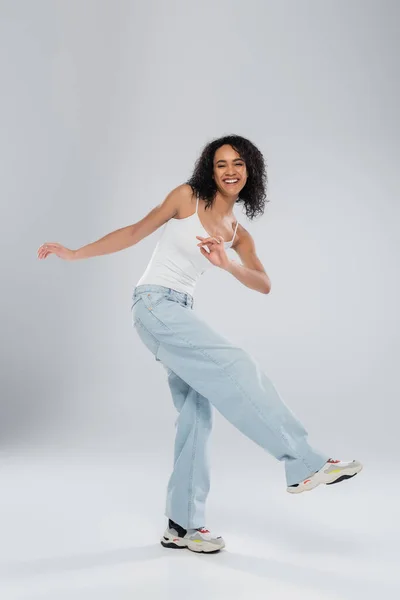 Повна довжина безтурботної афроамериканської жінки в білому баку зверху і джинси позують на сірому фоні — стокове фото
