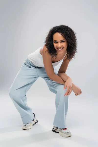 Volle Länge der überglücklichen afrikanisch-amerikanischen Frau in blauen Jeans und Turnschuhen posiert auf grauem Hintergrund — Stockfoto