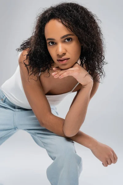 Curly african american woman in white tank top and blue jeans holding hand near chin and looking at camera isolated on grey — Stock Photo