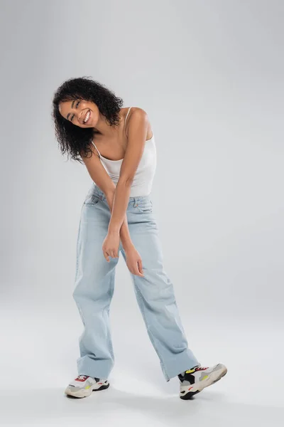 Volle Länge der fröhlichen afrikanisch-amerikanischen Frau in Turnschuhen und blauen Jeans posiert auf grauem Hintergrund — Stockfoto
