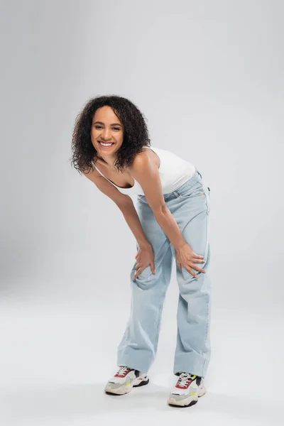 Pleine longueur de femme afro-américaine insouciante en jeans bleus souriant à la caméra sur fond gris — Photo de stock