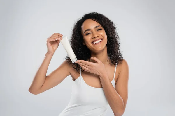 Cheerful african american woman in white tank top posing with tube of cosmetic cream isolated on grey — Stock Photo