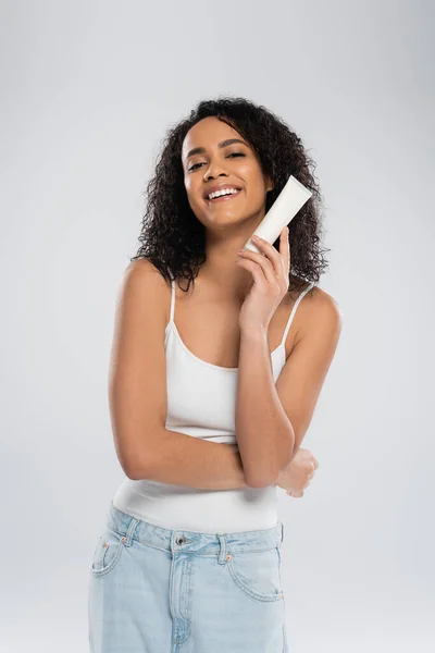 Mujer afroamericana complacida en camiseta blanca posando con tubo de crema cosmética aislada en gris - foto de stock