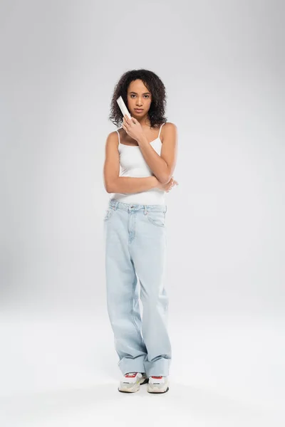 Comprimento total da mulher americana africana em top tanque branco e jeans azul segurando creme cosmético no fundo cinza — Fotografia de Stock
