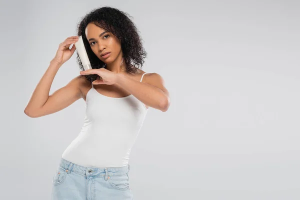Junge afrikanisch-amerikanische Frau in Tank Top und Blue Jeans zeigt kosmetische Creme isoliert auf grau — Stockfoto