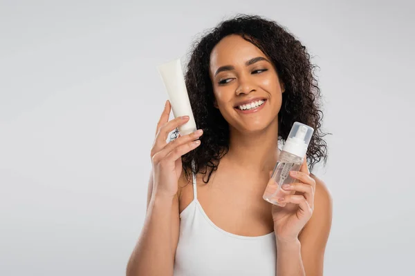 Heureuse femme afro-américaine tenant crème cosmétique et mousse visage isolée sur gris — Photo de stock