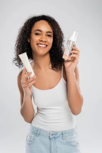 Brune afro-américaine en débardeur blanc tenant crème cosmétique et mousse visage isolée sur gris — Photo de stock