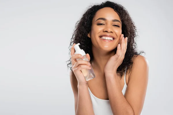 Mujer afro-americana feliz con pelo moreno rizado aplicando espuma facial aislada en gris - foto de stock