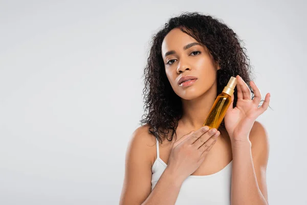 Jolie femme américaine africaine aux cheveux bruns bouclés tenant de l'huile cosmétique isolée sur gris — Photo de stock