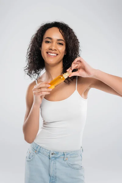 Aufgeregte afrikanisch-amerikanische Frau hält eine Flasche Kosmetiköl in der Hand, während sie isoliert auf grau in die Kamera blickt — Stockfoto
