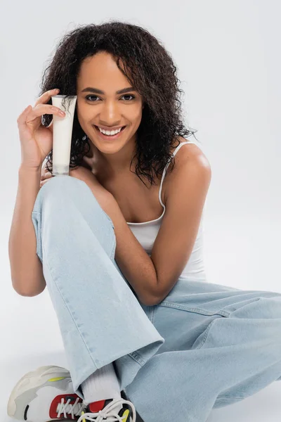 Усміхнена афроамериканка тримає трубку косметичного крему, сидячи на сірому фоні — стокове фото