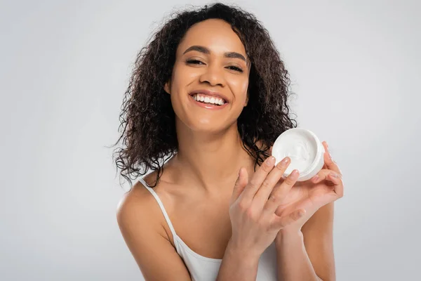 Young and happy african american woman with perfect skin touching cosmetic cream isolated on grey — Stock Photo