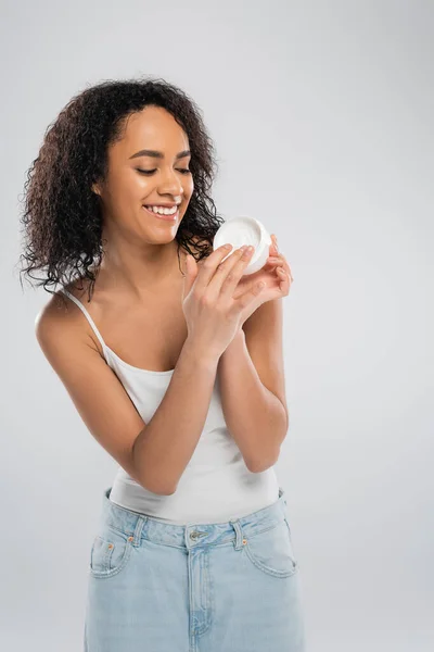 Mujer afroamericana complacida en camiseta sin mangas mirando contenedor de crema cosmética aislado en gris - foto de stock