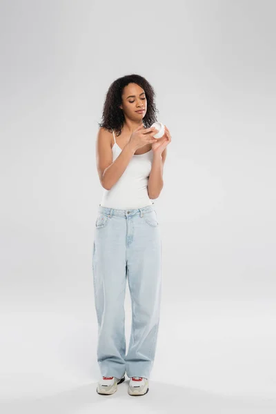 Longitud completa de la mujer afroamericana en pantalones vaqueros y camiseta mirando crema cosmética sobre fondo gris - foto de stock