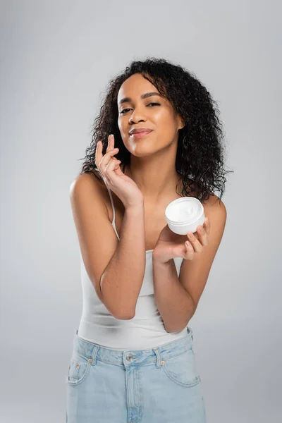 Encantadora mulher afro-americana olhando para a câmera e aplicando creme cosmético isolado no cinza — Fotografia de Stock