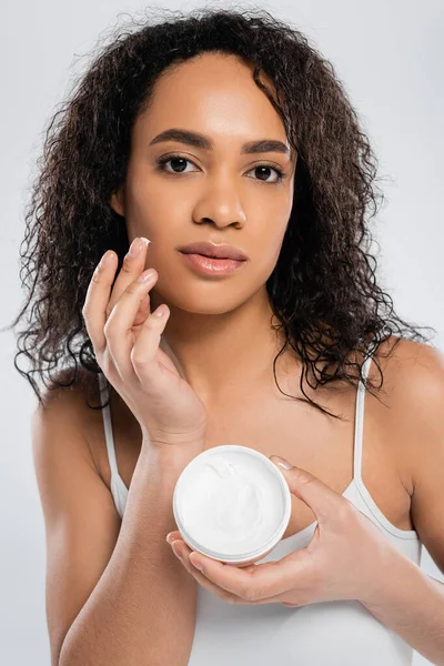 Mujer afroamericana con pelo moreno rizado aplicando crema facial aislada sobre gris - foto de stock