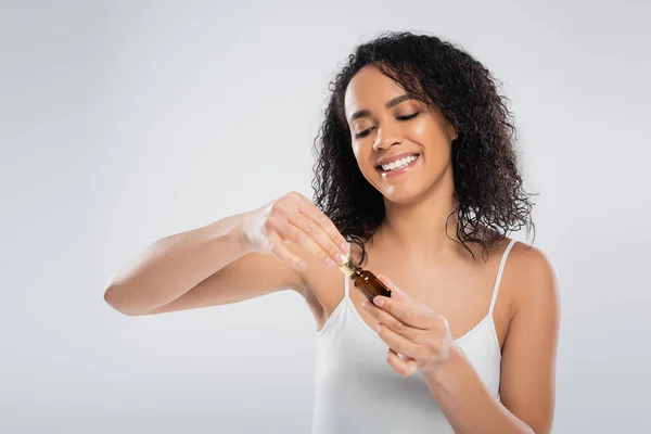Brünette afrikanisch-amerikanische Frau in weißem Tank Top Öffnung kosmetische Serum isoliert auf grau — Stockfoto