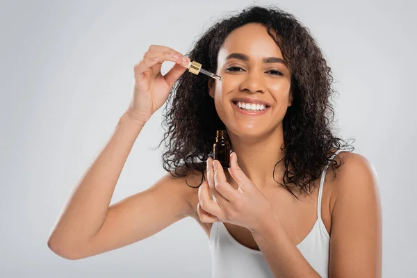 Freudige afrikanisch-amerikanische Frau Anwendung kosmetischen Serums auf perfekte Gesicht isoliert auf grau — Stockfoto