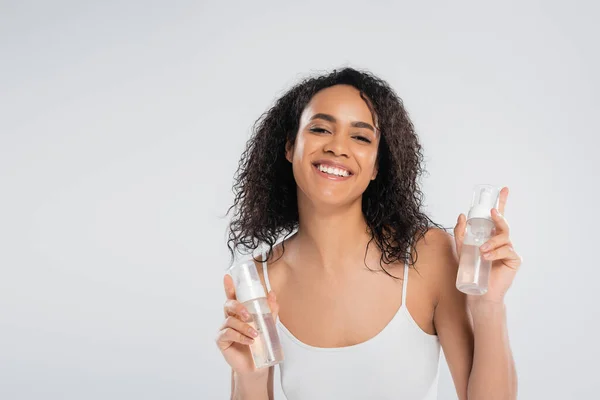 Joven mujer afroamericana posando con botellas de espuma facial mientras sonríe a la cámara aislada en gris - foto de stock