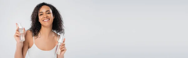 Felice donna afroamericana in posa con bottiglie di schiuma facciale mentre guarda la fotocamera isolata su grigio, banner — Foto stock