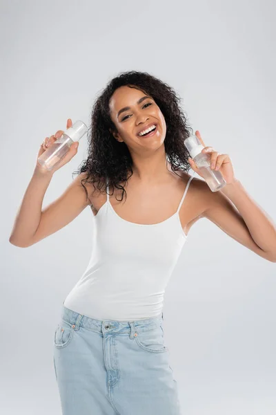 Overjoyed african american woman in white tank top posing with bottles of face foam isolated on grey — Stock Photo