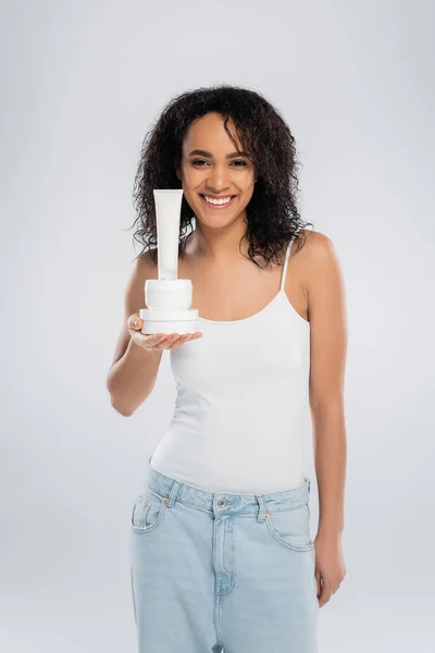 Joyful african american woman in tank top showing containers with cosmetic cream isolated on grey — Stock Photo