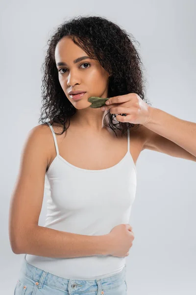 Young african american woman in white tank top using face scraper isolated on grey — Stock Photo