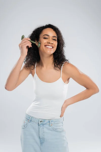Mujer afroamericana feliz en camiseta blanca de pie con la mano en la cadera y el uso de rodillo de jade aislado en gris — Stock Photo