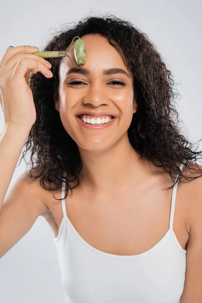 Mulher americana africana alegre fazendo massagem facial com rolo de jade isolado em cinza — Fotografia de Stock