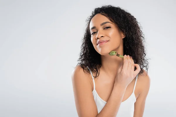 Curly african american woman using jade roller while smiling at camera isolated on grey — Stock Photo